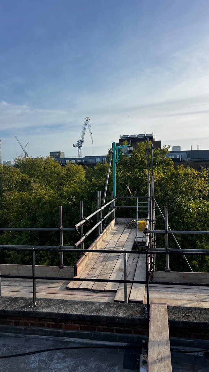 Armstrong Scaffolding boarded roof level bridge with an electric hoist mounted on a beam, looking out over the plane trees of Berkeley Square