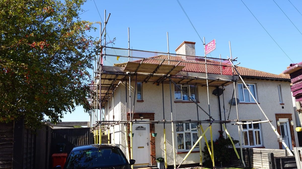 Boarded scaffold lift at eaves level across the front of a house in west London, one of twenty identical houses on the same roofing contract, with a mature tree at the boundary alongside