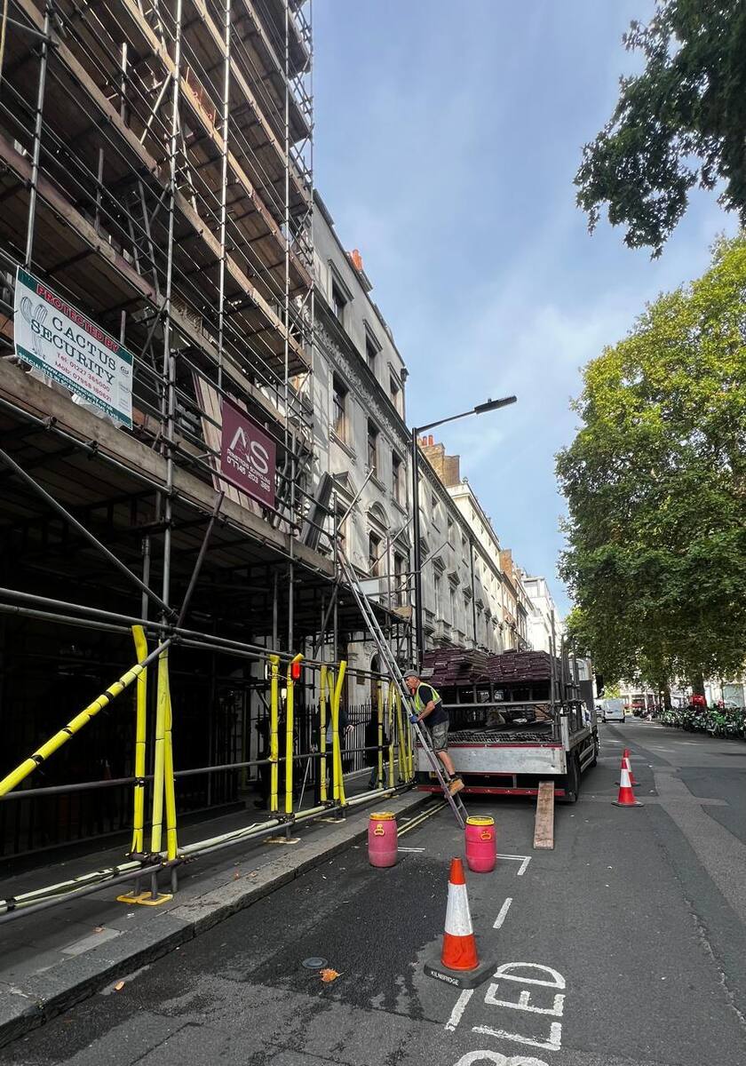 Armstrong Scaffolding crew unloading scaffold boards from a flatbed lorry in the carriageway beside the pedestrian gantry on Berkeley Square, Mayfair